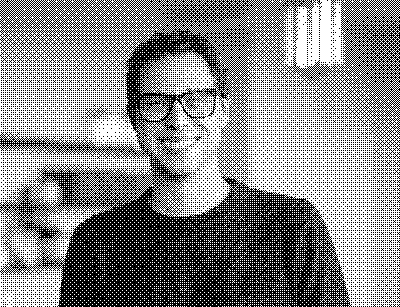 A smiling man with short, curly hair and black glasses, wearing a navy sweater in a softly lit room.